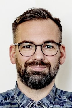 Mann mit dunklen Haaren, Brille, Vollbart und einem blau gemusterten Hemd